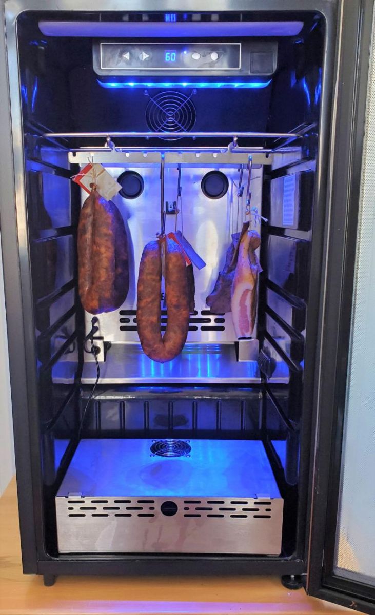 A modern SteakAger Pro 40 curing chamber set to 60 degrees, stocked with several hanging pieces of cured meats illuminated by a blue light, showcasing a clean and controlled environment for meat aging.