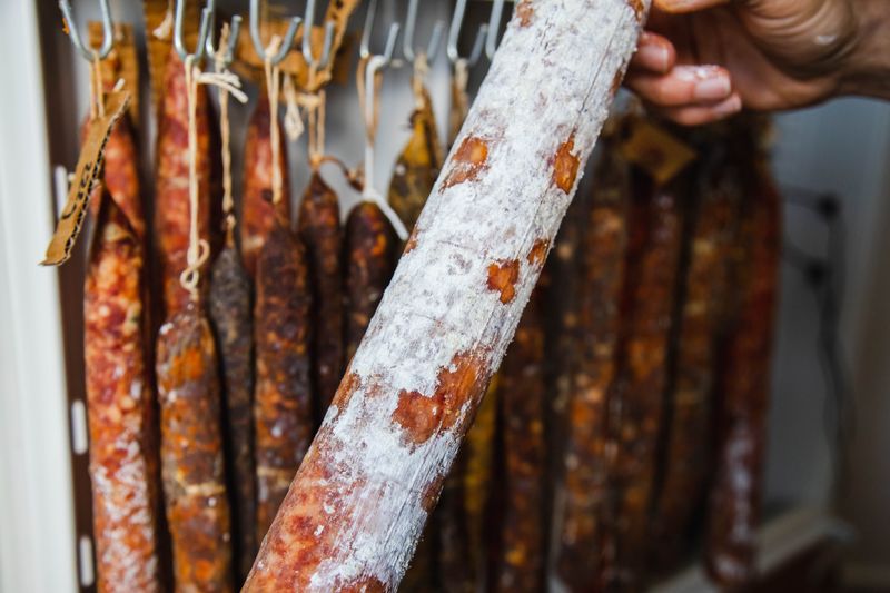 A closeup picture showcasing a person's hand holding a homemade salami, with a variety of other sausages hanging in the background, covered in white mould which is typical in the curing process.