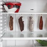 Various types of preserved meats hanging inside a refrigerator, exhibiting the process of aging or storage for flavor development.