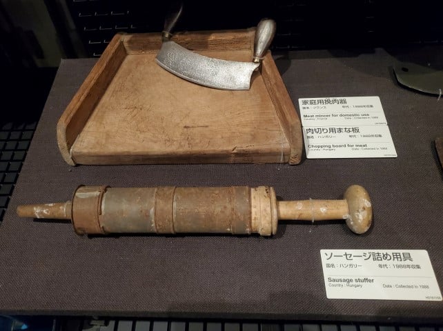 An antique sausage stuffer and meat cleaver displayed on a table next to an informational placard about making salami at home, showcasing culinary tools from a bygone era.