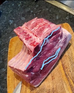 A large raw cut of beef with a bone section lies on a wooden cutting board, ready for preparation.