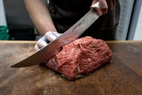 A chef wearing gloves skillfully prepares to grind a fresh, raw whole beef brisket on a wooden cutting board, aiming for economical cuts.
