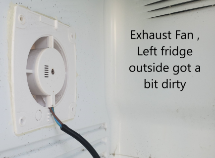 An exhaust fan mounted on the interior wall of a fridge's external compartment, showing signs of dirt and dust accumulation.