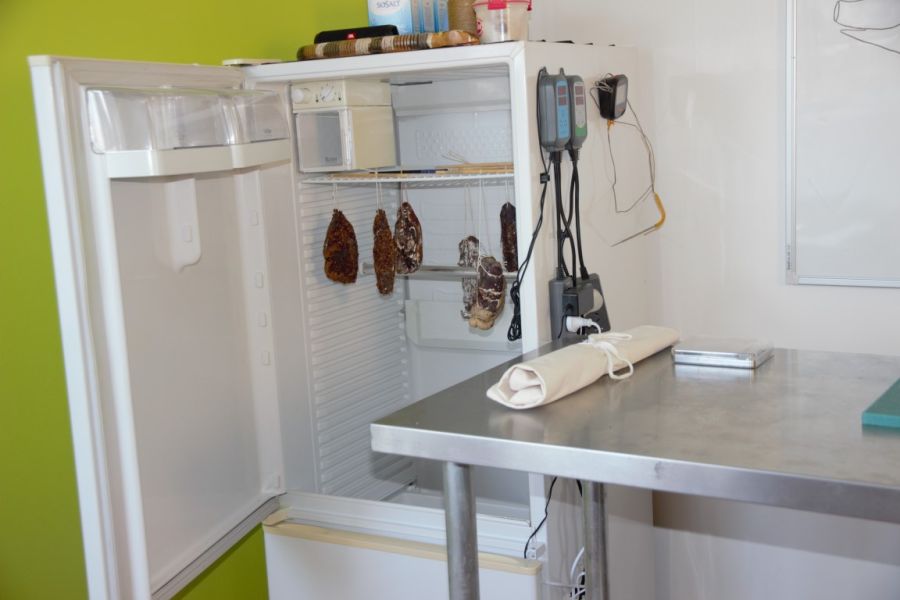 Open refrigerator, regulated by an INKBIRD ITC-308 temperature controller, with almost empty shelves featuring a few food items and a pair of sausages hanging inside, next to a kitchen counter