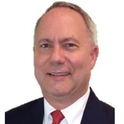 A professional headshot of a smiling man wearing a suit with a white shirt and red tie, holding craft cured meats.