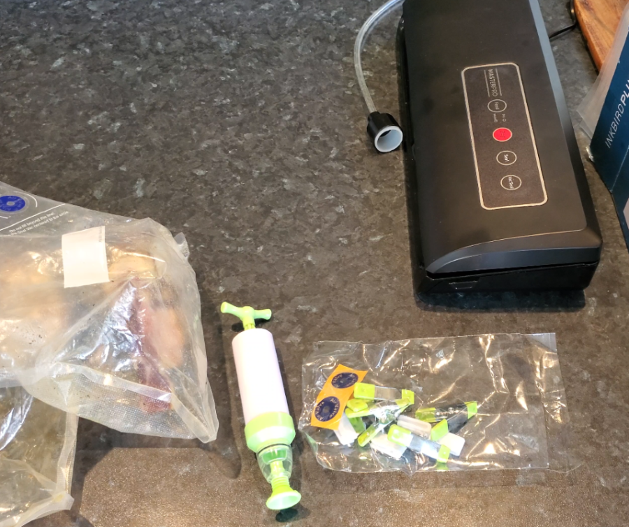 Vacuum sealing tools and accessories laid out on a kitchen countertop, including a vacuum sealer machine, a hand pump, plastic sealing bags for storing cured meat, and various vacuum seal valves.