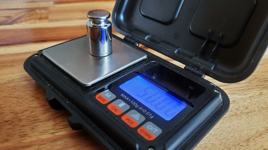 Close-up of a digital pocket scale, crucial equipment for equilibrium curing, with a metal calibration weight on top, displaying measurements on a blue backlit LCD screen.