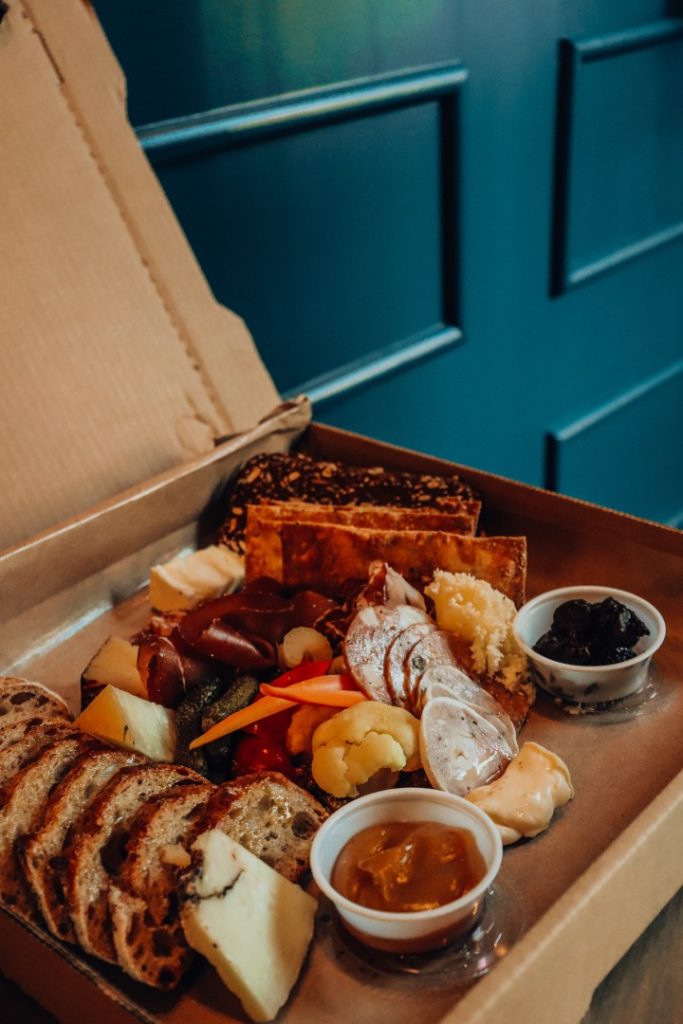 A gourmet charcuterie board, complete with an assortment of cured meats, cheeses, fruits, and condiments, served with slices of crusty bread and presented in a casual to-go box setting.