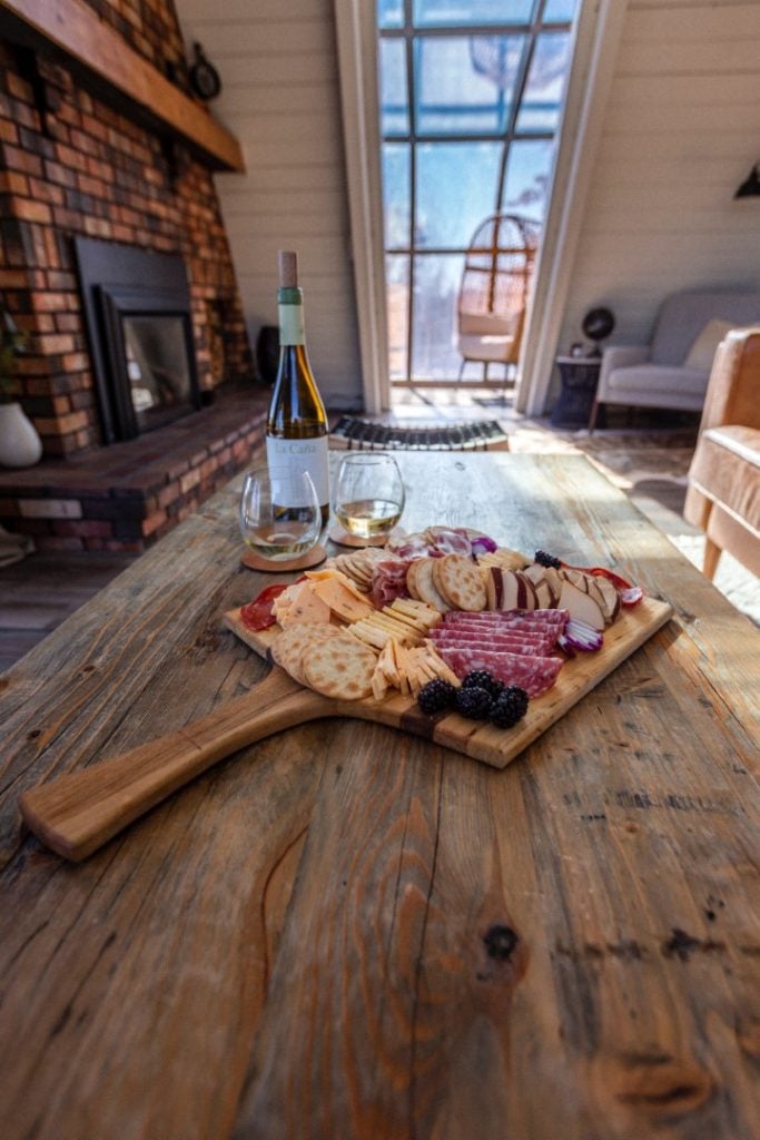 A cozy indoor setting featuring a charcuterie board laden with assorted cheeses, cured meat, and berries, perfectly paired with a bottle of wine and two glasses, with the inviting ambiance of a fireplace and