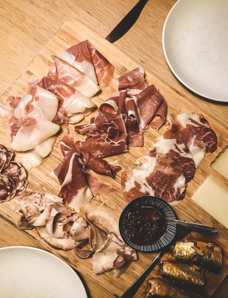 A savory Charcuterie Board essentials with an assortment of cured meats, cheese slices, and a small bowl of jam, arranged on a wooden surface for a delightful appetizer experience.