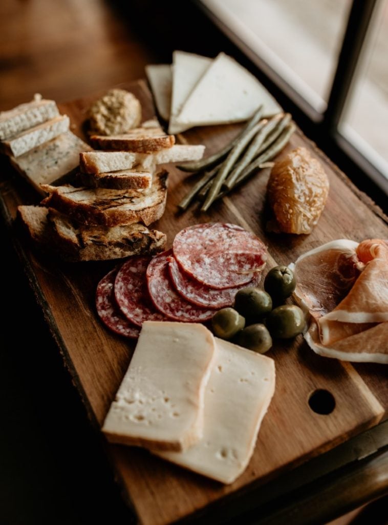 A rustic and appetising charcuterie board featuring an assortment of cheeses, slices of bread, cured meats, green olives, and pickles, artistically arranged on a wooden serving board. Discover