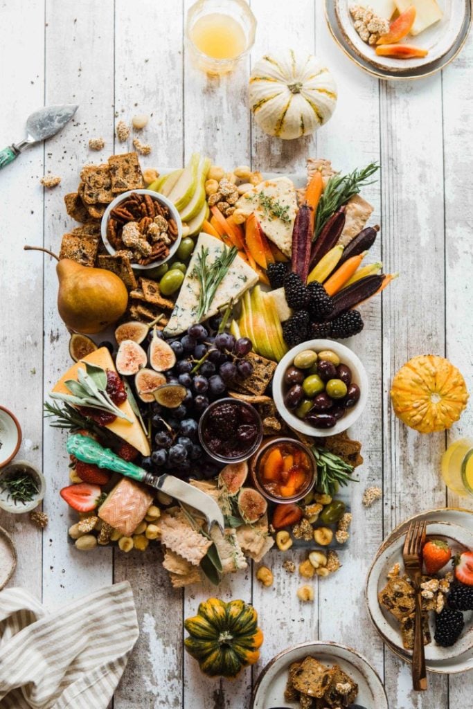 A festive and abundant autumnal charcuterie board spread with a variety of cheeses, cured meat, fruits, crackers, and nuts arranged on a rustic white wooden surface, flanked by mini pumpkins