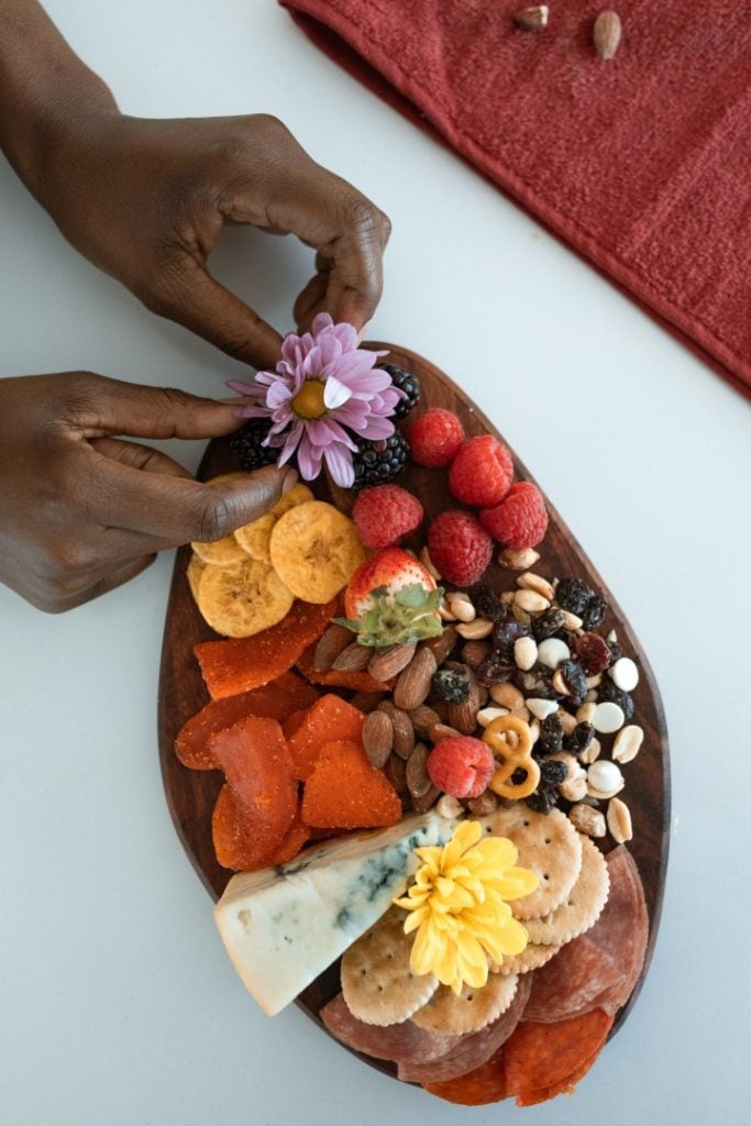 A person's hand delicately placing a flower on a colorful charcuterie board filled with an assortment of fruits, nuts, cheeses, and essential cured meats.