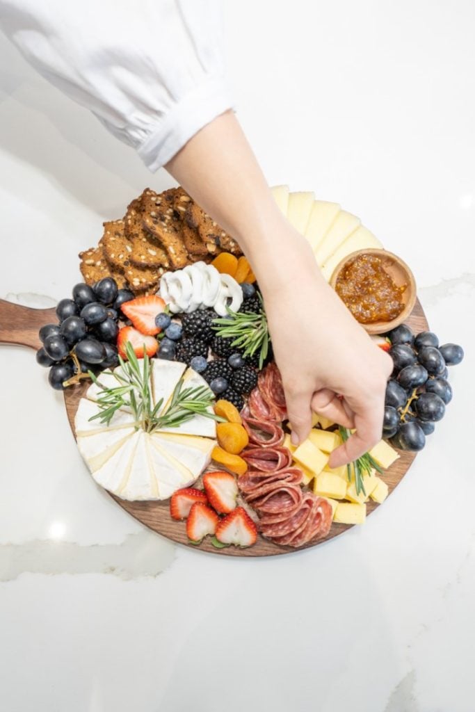 A person's hand arranging a colorful and artfully displayed charcuterie board with a variety of cheeses, fruits, cured meats, and condiments on a wooden platter.