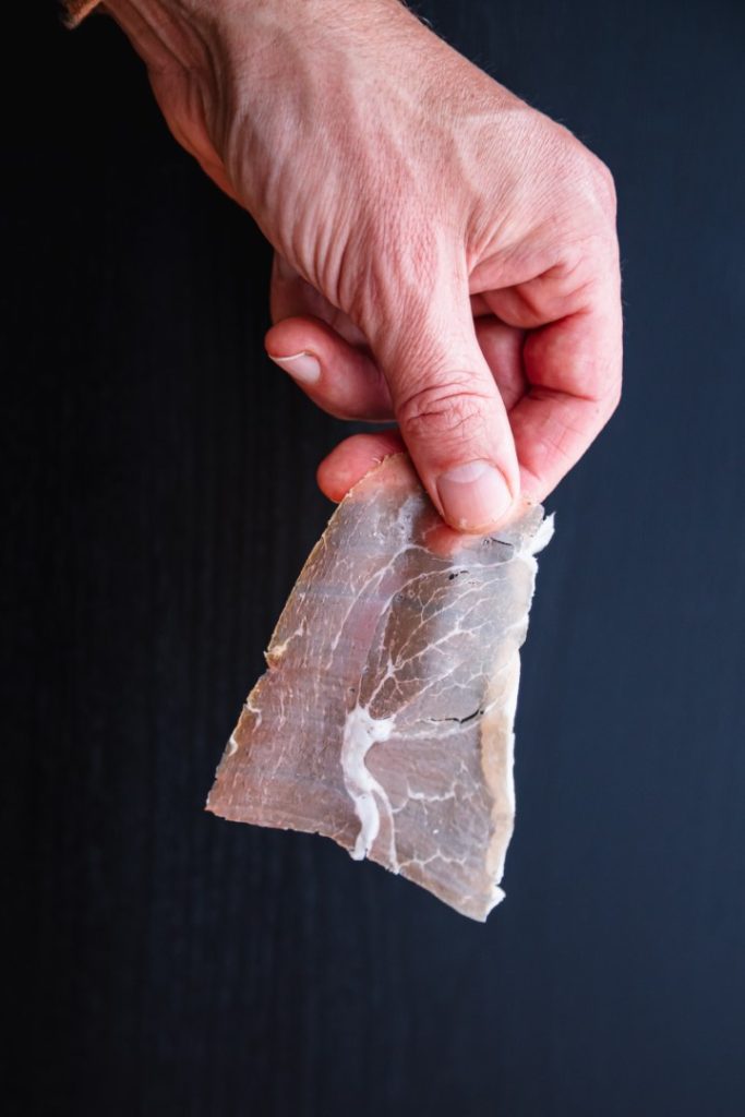 A hand holding a thin slice of cured meat against a dark background.