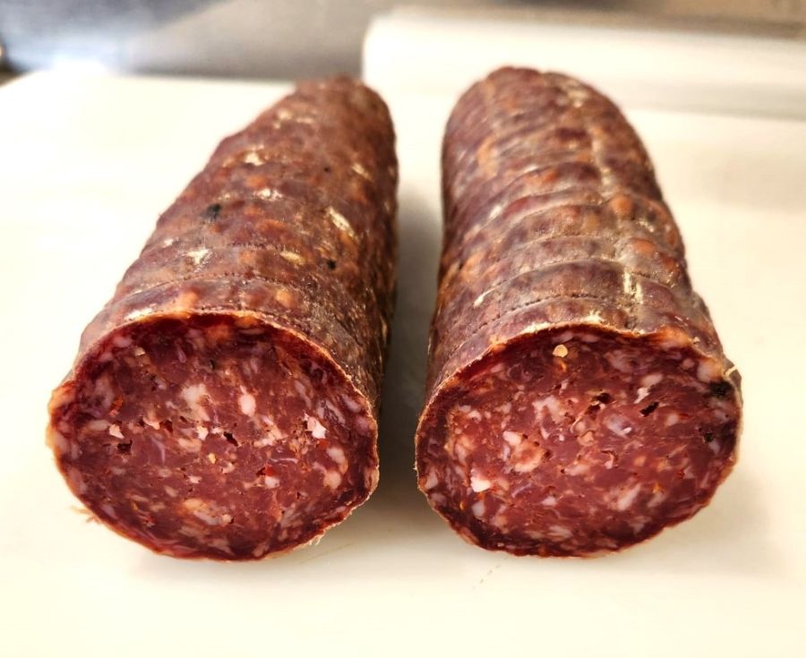 Two homemade salami sausages, one whole and one sliced at the end, showcasing a marbled interior with spices, displayed on a white surface.