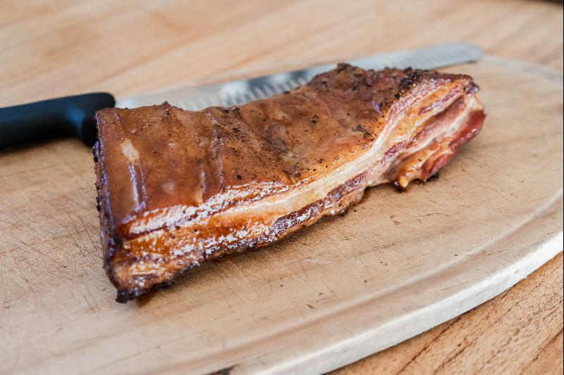 A succulent piece of grilled pork belly, now transformed into bacon, rests on a wooden cutting board next to a chef's knife, showcasing a perfectly crispy skin and juicy layers of meat and fat.