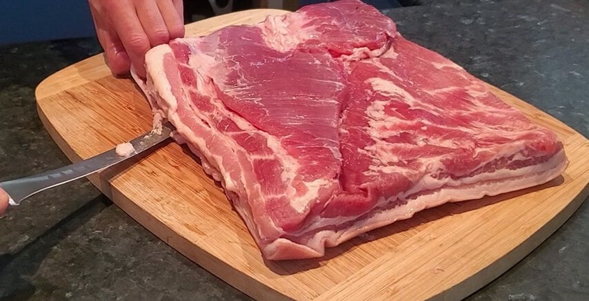 A hand holding a knife trims fat from a large piece of raw, skin-on bacon on a wooden cutting board.