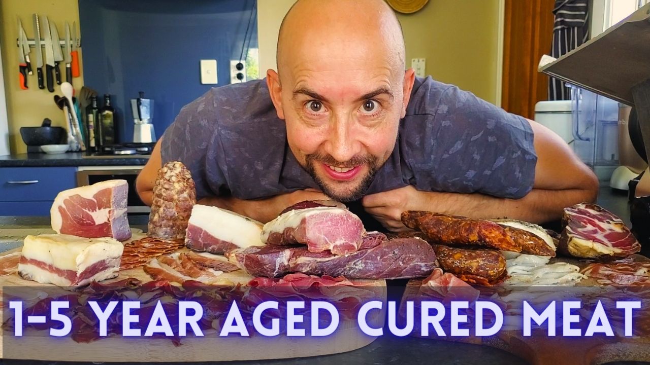 A person with a shaved head is leaning forward on a kitchen counter, surrounded by various types of aged cured meats. The text "1-5 Year Aged Cured Meat" is displayed prominently at the bottom of the image. Kitchen utensils and appliances are visible in the background, perfect for trying out cured meat recipes.