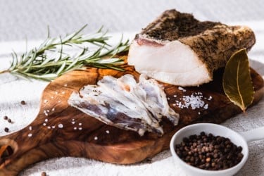 picture of salt cured fat on a board with rosemary and peppercorns around it.