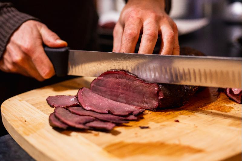 A person slicing a piece of cooked meat with a large knife on a wooden cutting board. The meat appears medium-rare with a reddish-pink center and darker outer edges, typical of hot smoking. The person is holding the knife with one hand and steadying the meat with the other in this beginner's guide technique.