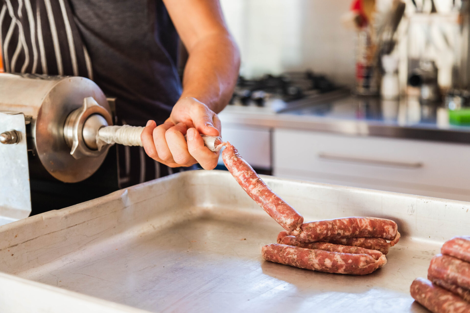 A person is seen using a sausage stuffer to fill sausage casings over a large metal tray in a kitchen. The tray contains several links of cured meat. The individual is wearing a dark shirt and a striped apron, with the kitchen counter visible in the background.