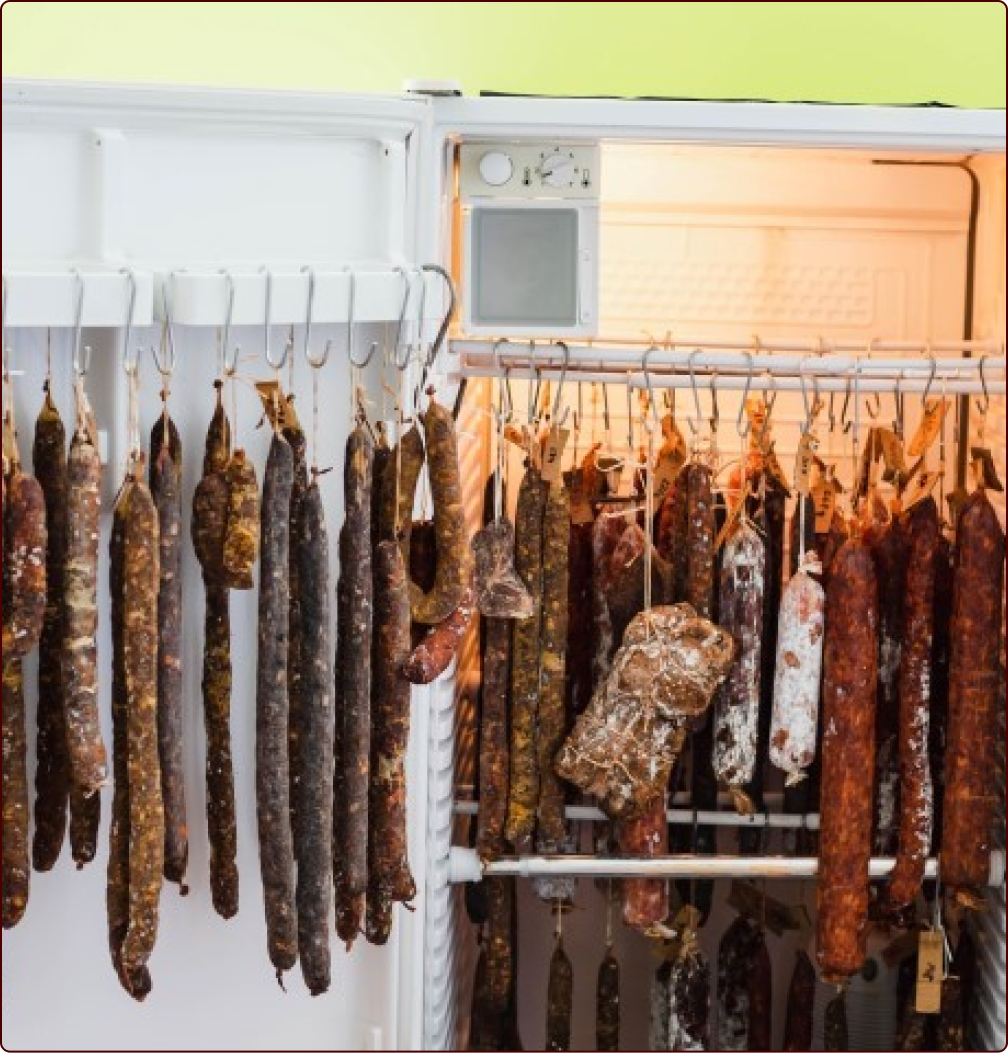 An open refrigerator is shown, filled with numerous hanging dried sausages and cured meats of various types and colors. The sausages are hung on hooks in both the main compartment and the door of the refrigerator. The fridge light casts a warm glow inside.